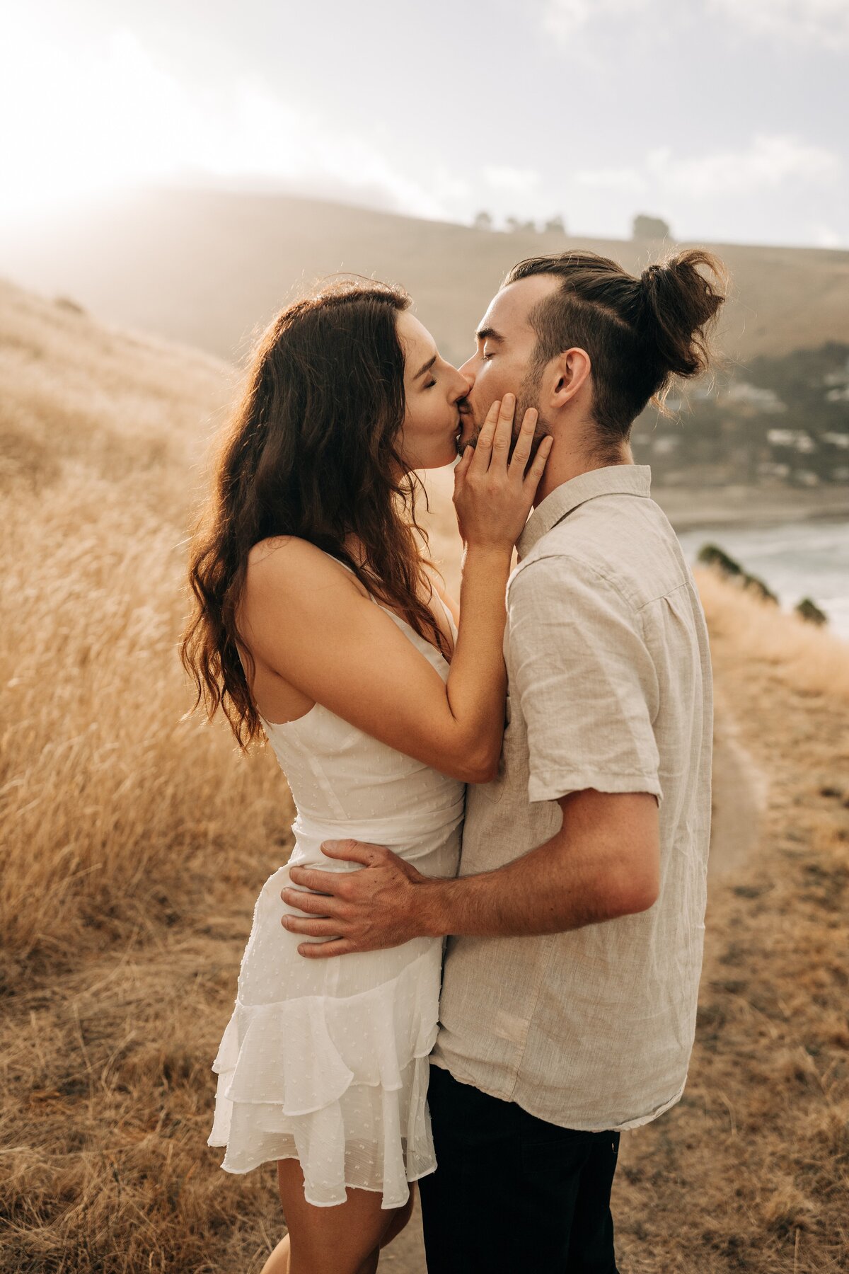 engaged couple in white dress and oat linen shirt kiss at taylors mistake in christchurch at sunset
