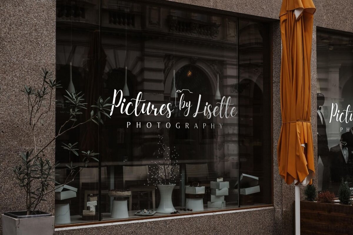 Large storefront window with white vinyl lettering “Pictures by Lisette Photography,” reflecting a city street and cafe setting.