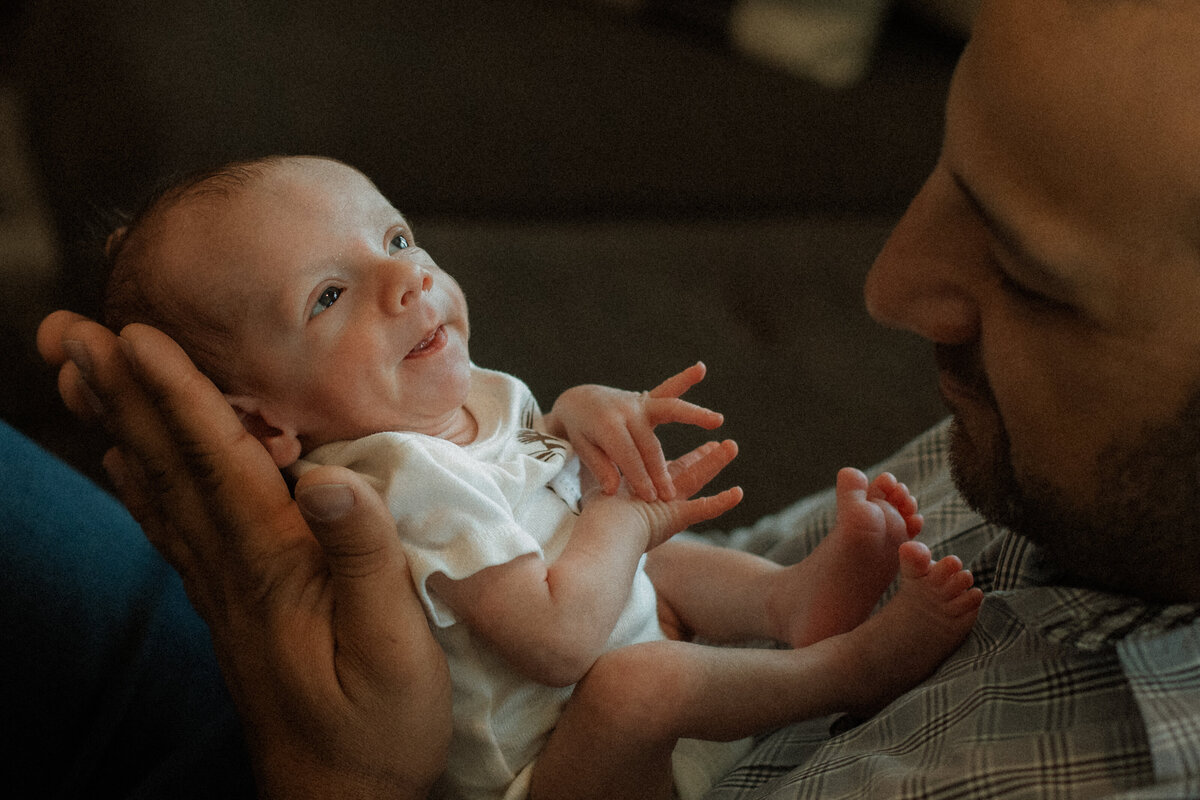 Newborn sitting on dad's chest while looking up bathed in window light.
