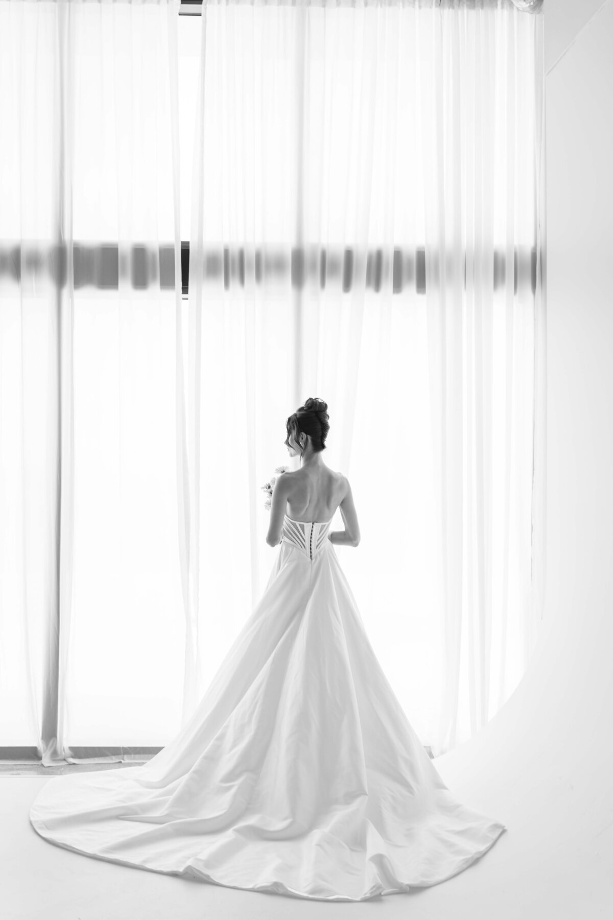 black and white photo of a bride facing a big window, and giving her back to the camera