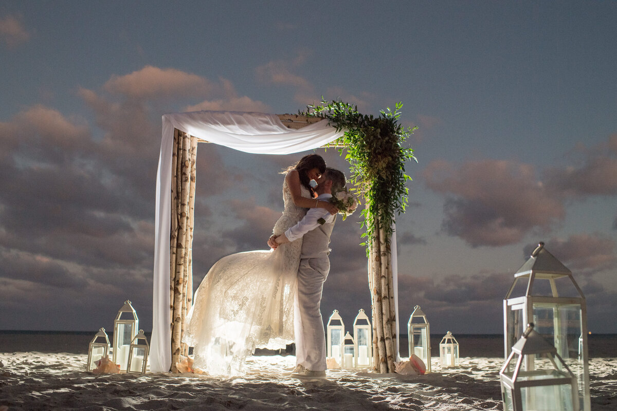 A groom dips his bride under a rustic beach wedding arch at sunset, captured by Rodrigo Varela Photography.
