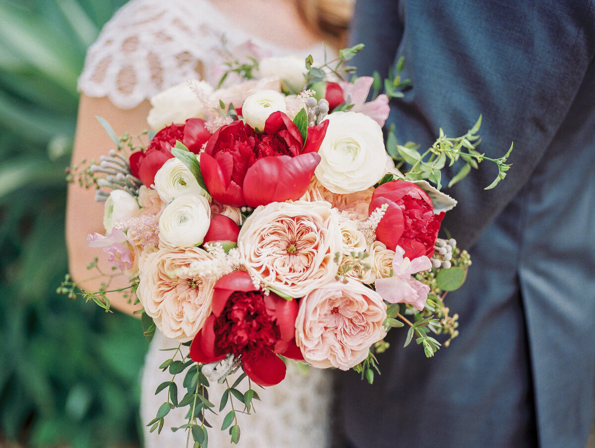 Luxury bridal bouquet featuring coral garden roses, red peonies, white ranunculus, and blush David Austin roses. Romantic wedding florals with cascading greenery and delicate textures. Fine art wedding photography.