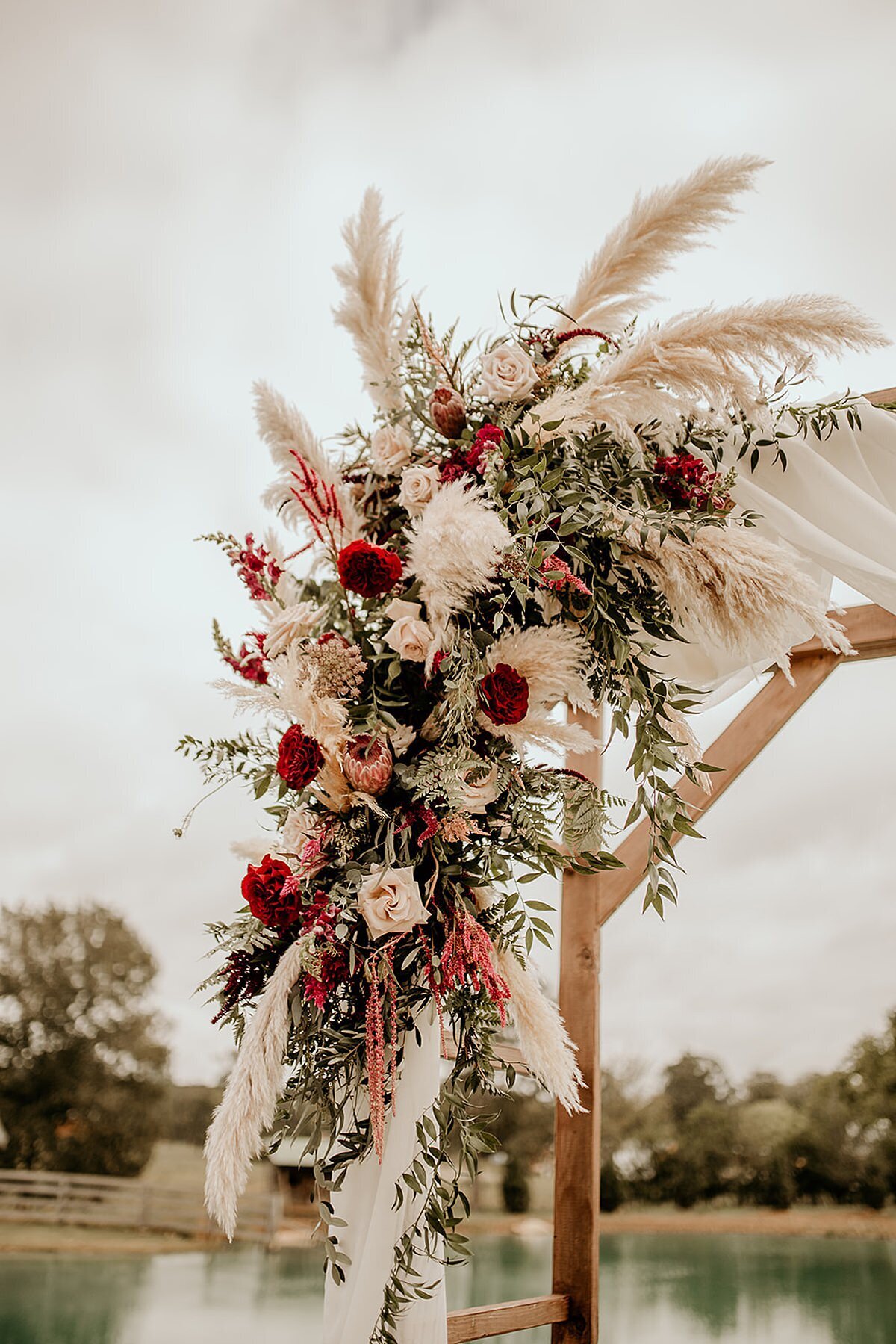 Wood ceremony arbor at Steel Magnolia Barn decorated with a boho floral spray of pampas grass, Italian ruscus, protea, burgundy carnations, blush roses, and pink rice flower along a pond.