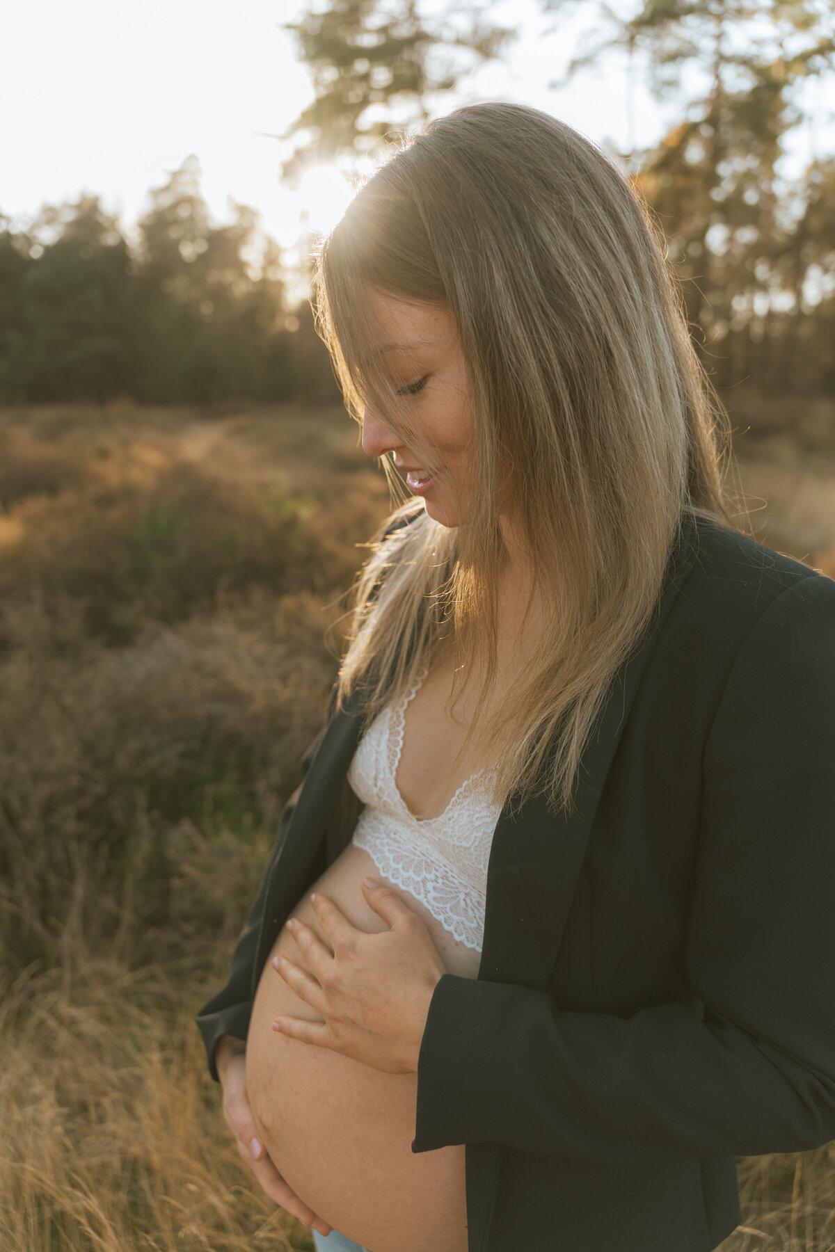 Zwangere vrouw staat met blote buik en kostuumvest in veld met ondergaande zon.