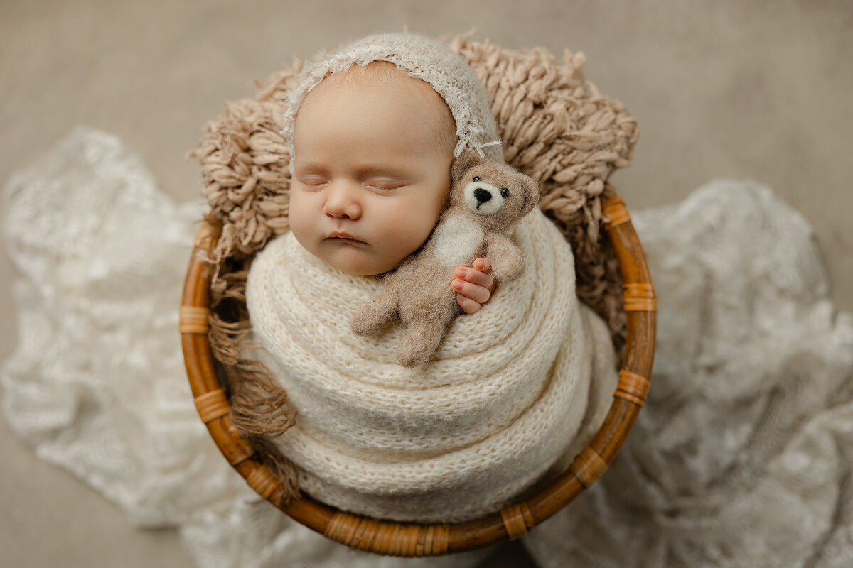 Sleeping baby girl swaddled in cozy neutrals with a delicate lace bonnet, cuddling a felted bear in a warm-toned studio setup. Photographed by Inland Empire newborn photographer Kaitlyn Dawn.