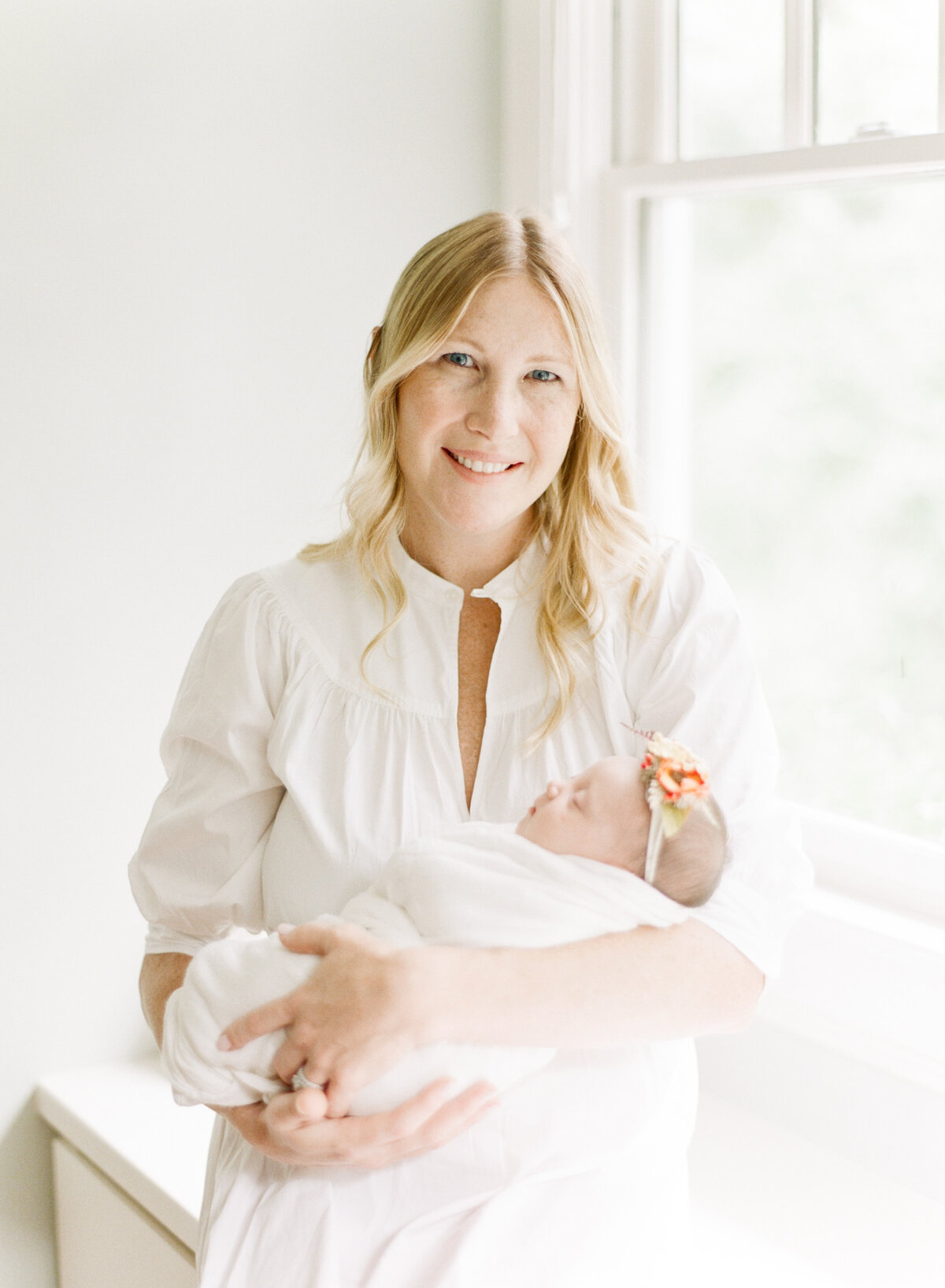 Mom smiles for the camera while holding her newborn as she's bathed in light from a nearby window. Image by Raleigh newborn photographer A.J. Dunlap Photography.