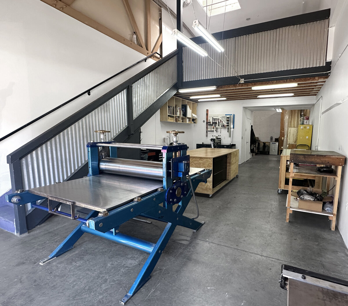 A spacious printmaking studio featuring a large blue press machine, work tables, and an organized tool wall.