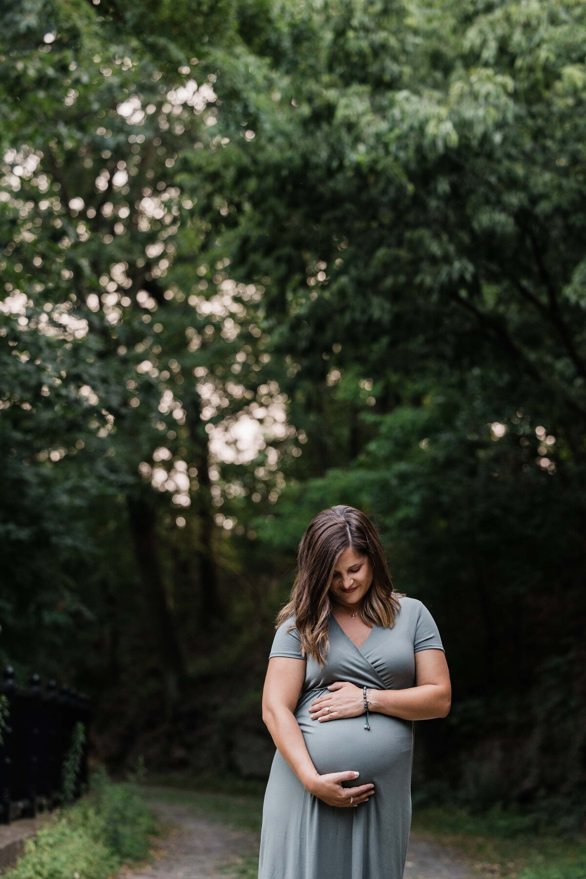 A pregnant woman smiling down at her belly while standing on a path with greenery in the background, captured by a Pittsburgh maternity photographer.