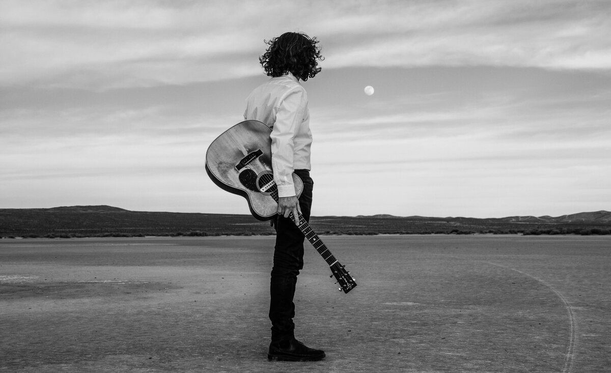Country musician portrait black and white Noah Zacharin holding guitar wearing white dress shirt black jeans looking out towards moon desert background