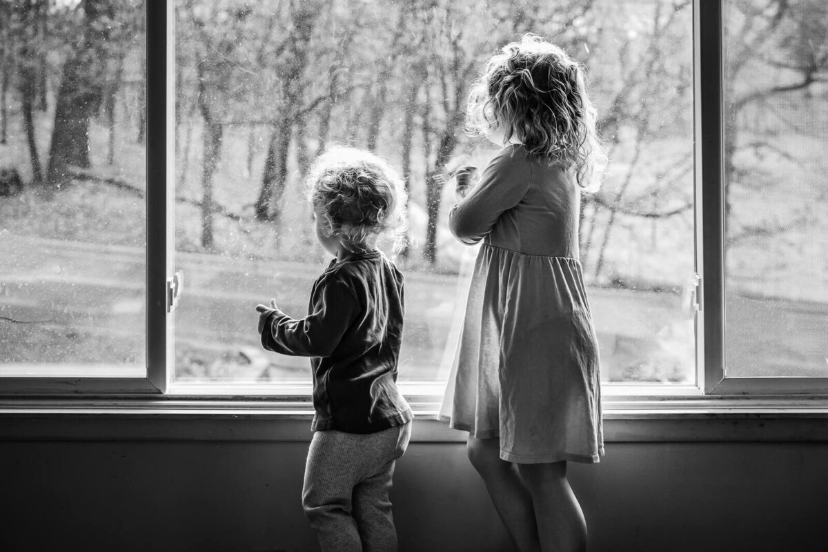 A black and white image of a young girl and her little brother looking out their front window.