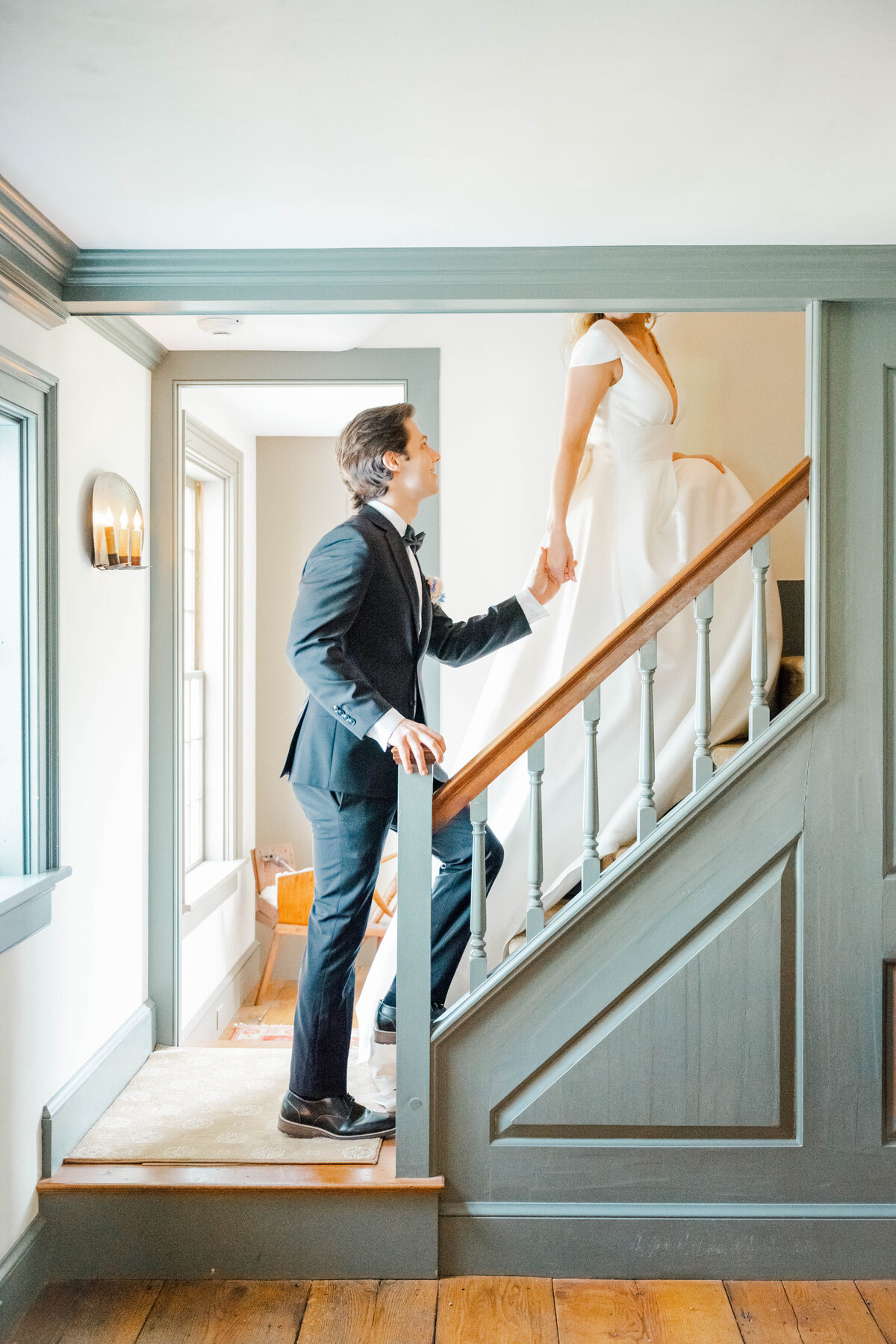Rhode Island Wedding Photographers | A couple on a staircase, the groom in a black tuxedo holding the hand of the bride in a white gown. Bright natural light fills the elegant setting.