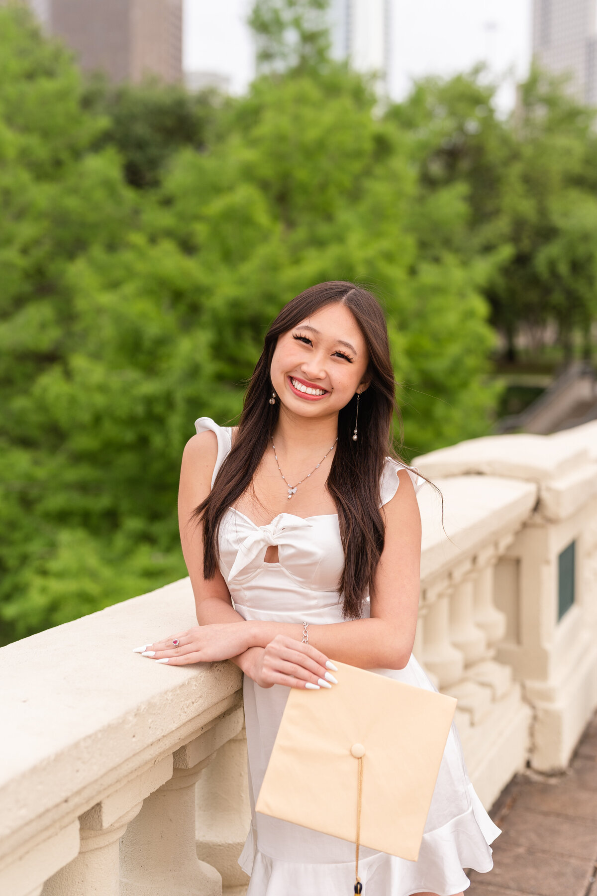 Houston high school senior girl leaning against white bridge railing and holding grad cap and smiling in Downtown Houston