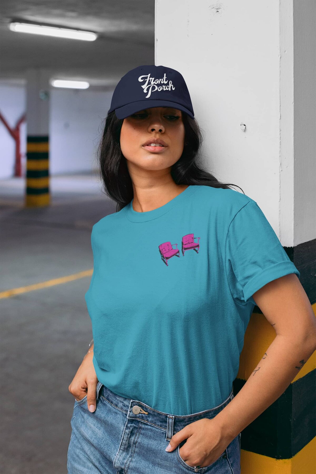 A woman standing in a parking garage wearing a navy baseball cap with "Front Porch" embroidered on the front, and a teal t-shirt with a pair of pink chairs on the left chest