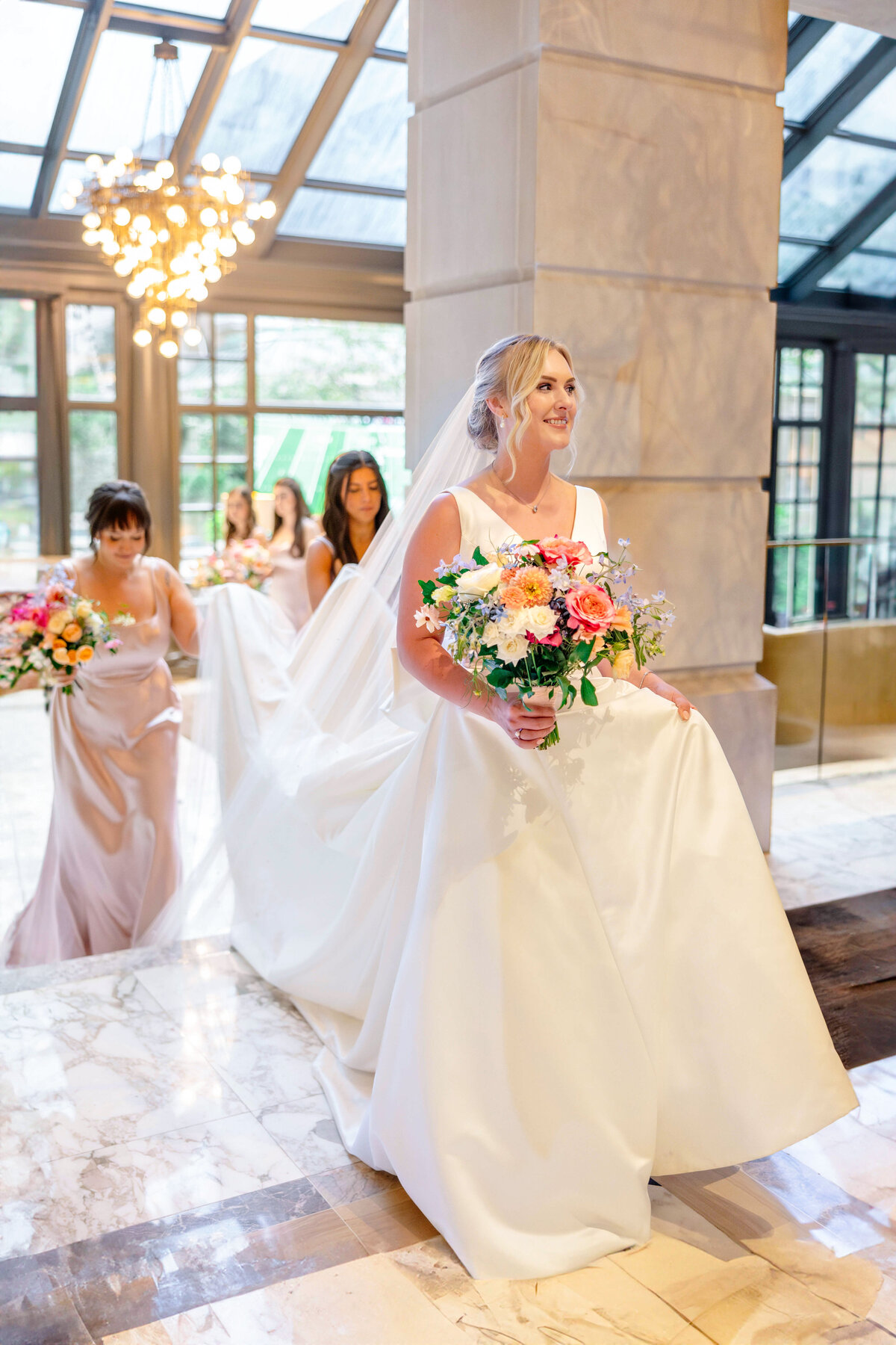 A bride in a white gown walks indoors, holding a colorful bouquet. Three bridesmaids in light pink dresses follow her, each carrying bouquets. The setting features marble floors and large windows with hanging lights.