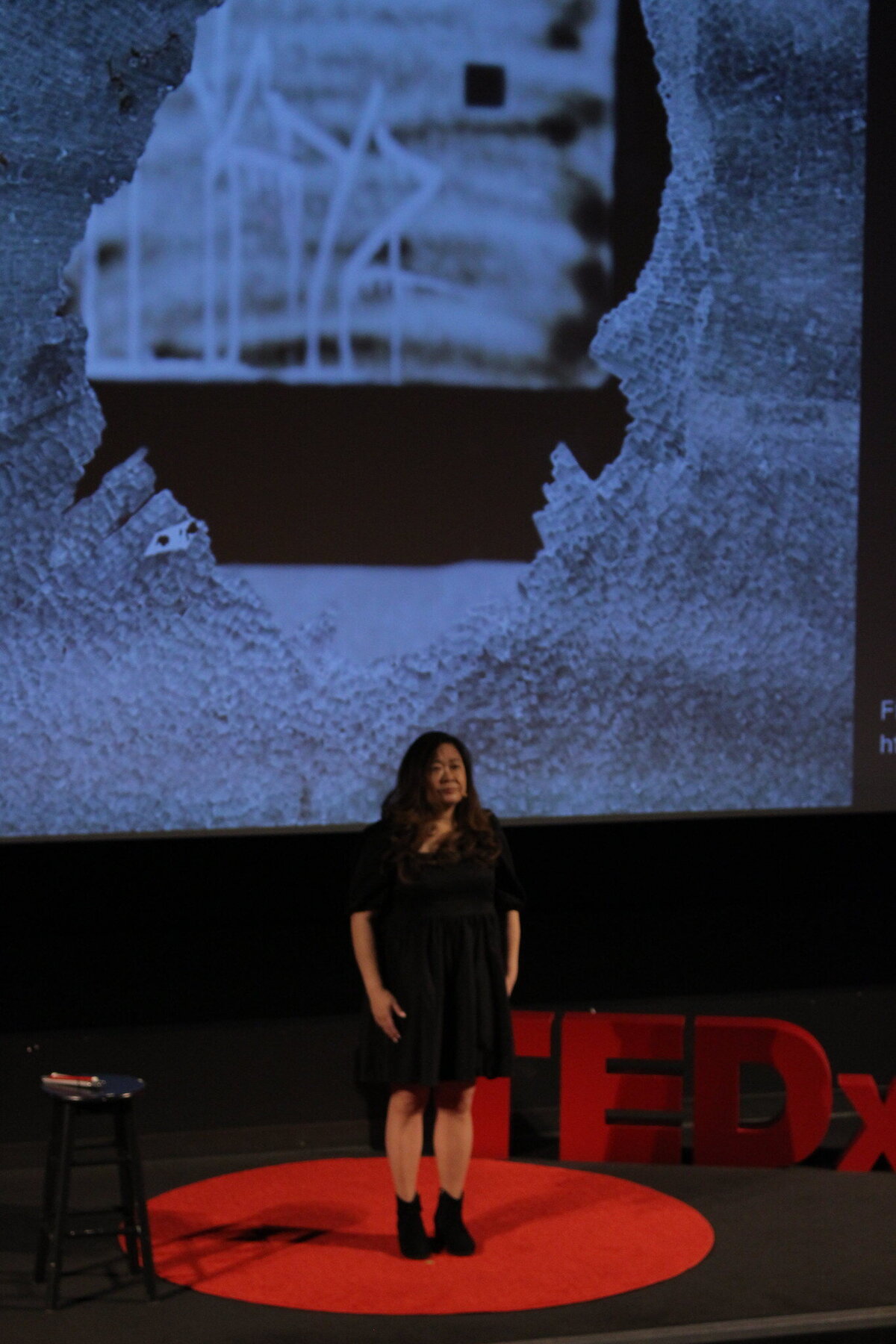 Anne standing at centre TEDxSFU stage with screen image of a window broken demonstrated by a hole through it