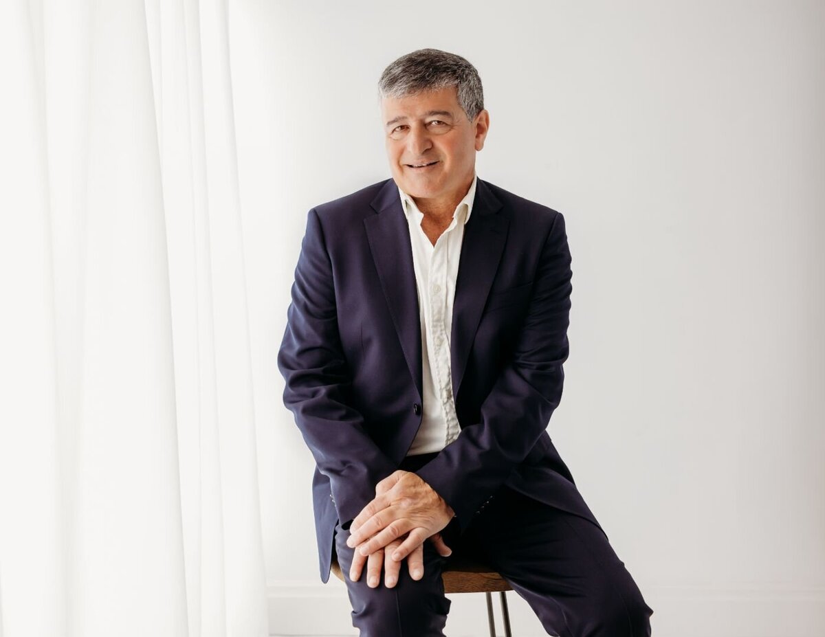 Grey-haired man in blue suit seated on stool, hands clasped on lap, pleasant smile. Photo by Ruaa Hamdan Photography