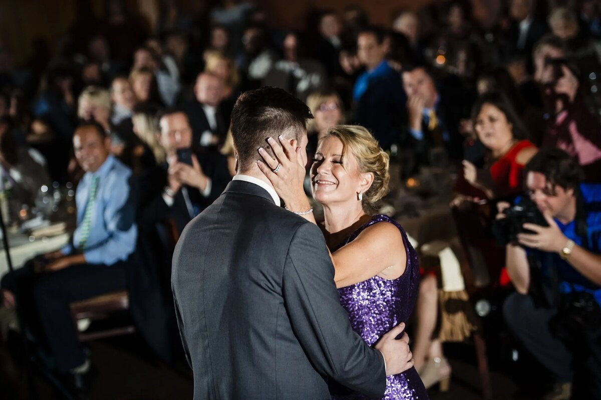 A heartfelt dance between a man in a suit and a woman in a purple dress, surrounded by warmly smiling wedding guests, encapsulating a tender moment.