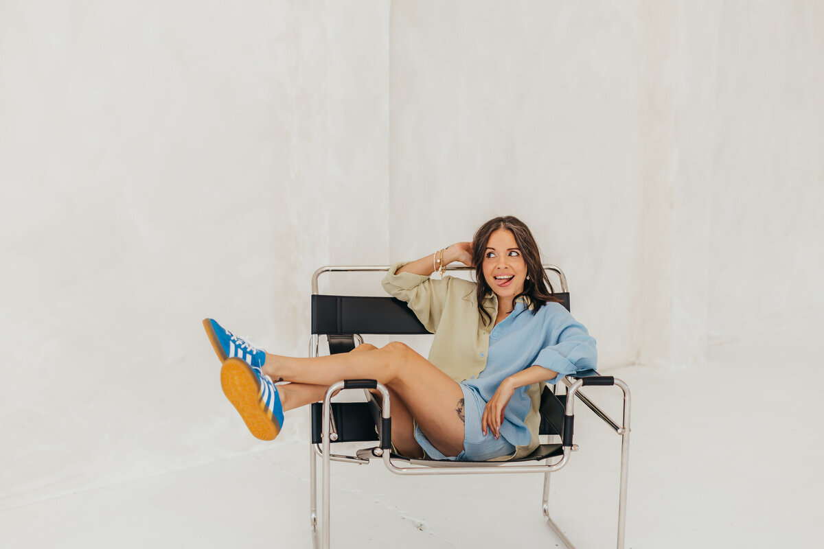 Sitting sideways on a studio chair with legs draped over the armrest, tongue out, and smiling- bringing big personality and playful energy to the creative space as a bold web designer.