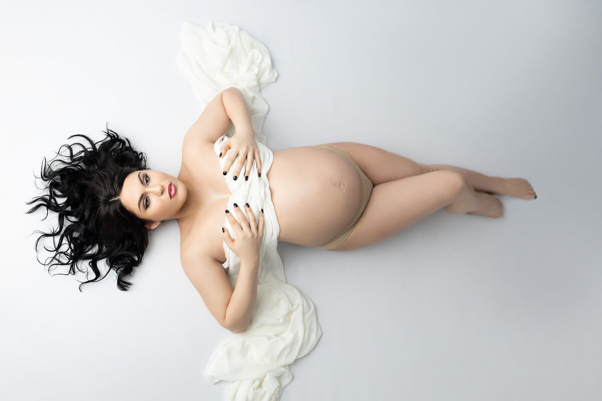 A stunning maternity photograph featuring a woman lying on a white backdrop, draped in soft white fabric that gently wraps around her body. Her dark hair cascades beautifully, enhancing the elegant simplicity of the scene.