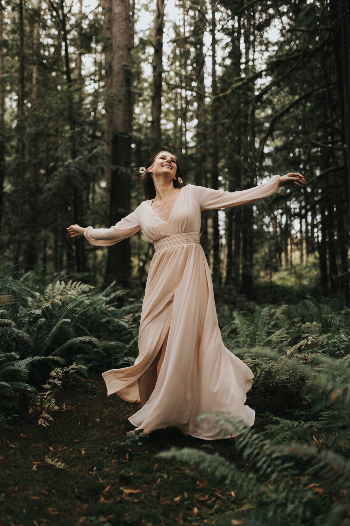 Woman stands in dark, lush forest wearing a cream-colored flowy dress with arms outstretched beside her.