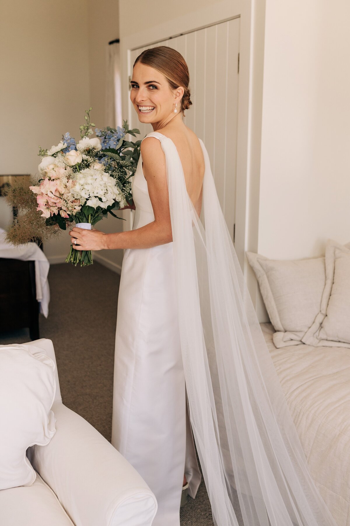bride in emilia wickstead dress real wedding with cape holding bouquet in hotel bedroom blenheim new zealand smiling summer