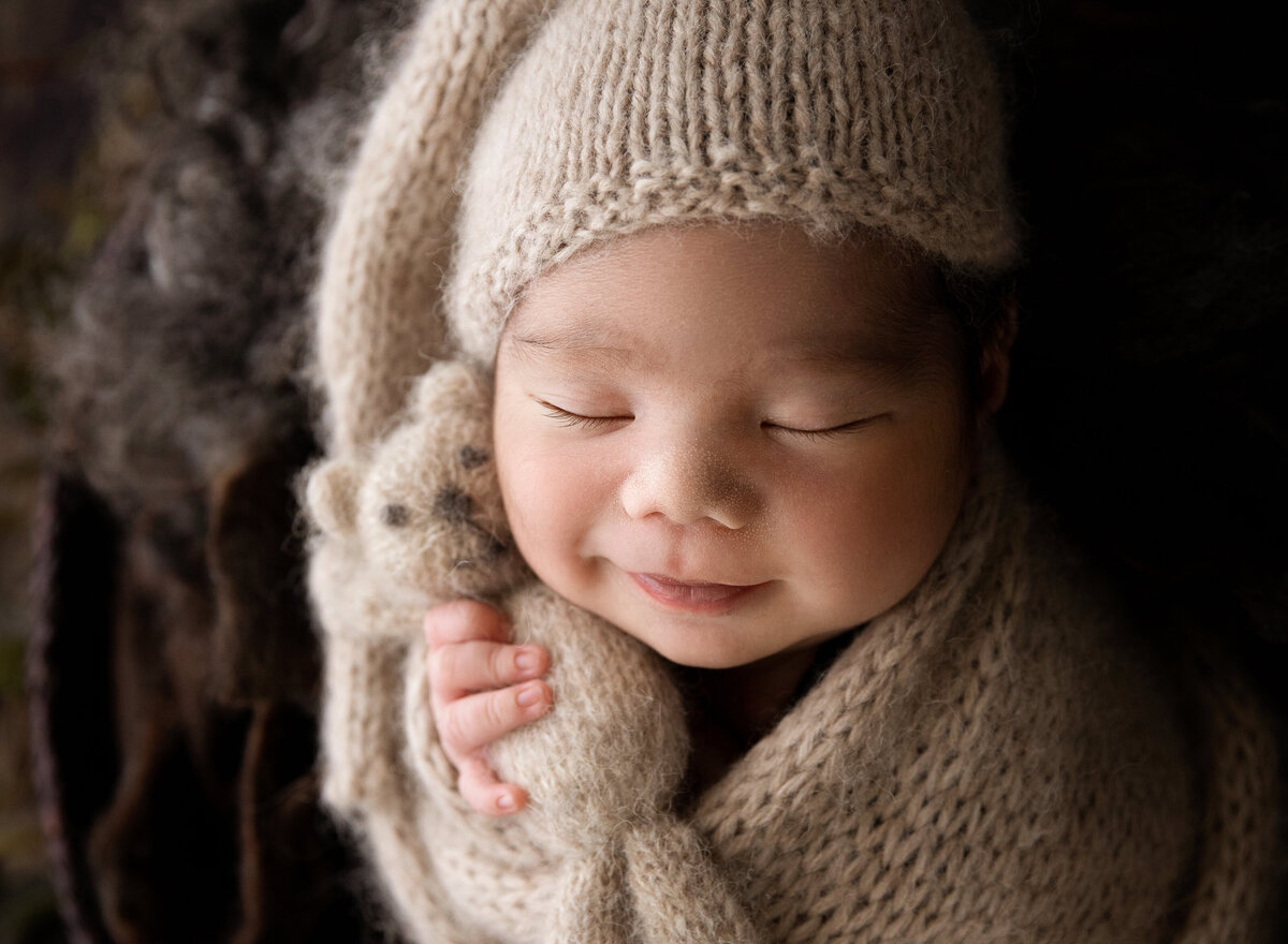 newborn baby boy snuggled in cream sleepy hat and wrap holding a soft teddy and smiling