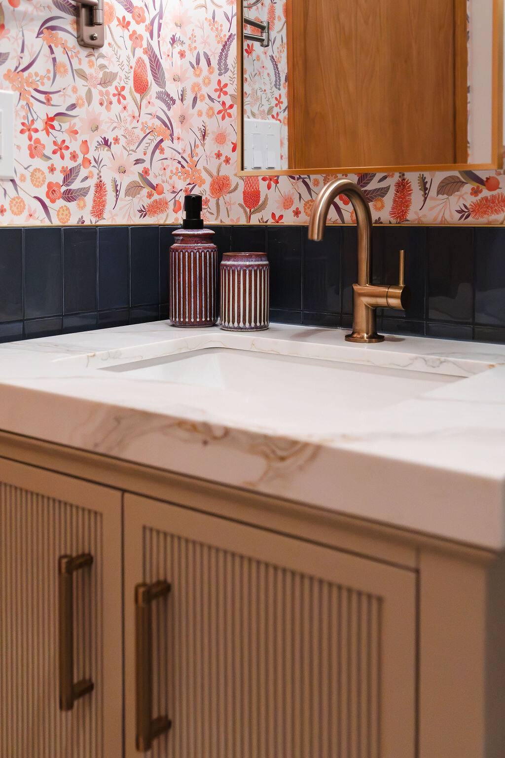 A close-up of a bathroom vanity featuring a white Calacatta Gold Quartz countertop with subtle veining. A rectangular undermount sink is fitted with a modern brass gooseneck faucet. Several decorative items sit on the countertop: a tall, ribbed brown glass bottle with a black pump, a shorter, ribbed metallic brown tumbler, and a small blue glass bottle with a cork stopper. The backsplash is made of dark blue vertical tiles. The wall above the tiles is covered with a vibrant floral patterned wallpaper in shades of pink, orange, and white. A partially visible wooden-framed mirror hangs above the sink. Light switches are visible on the wall to the left. A glimpse of a white towel with tassels is on the right.