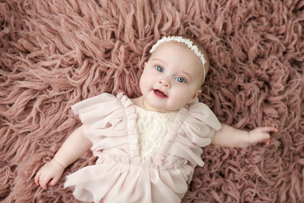 Smiling baby girl milestone portrait with soft pink textures and delicate ruffles—pure sweetness.