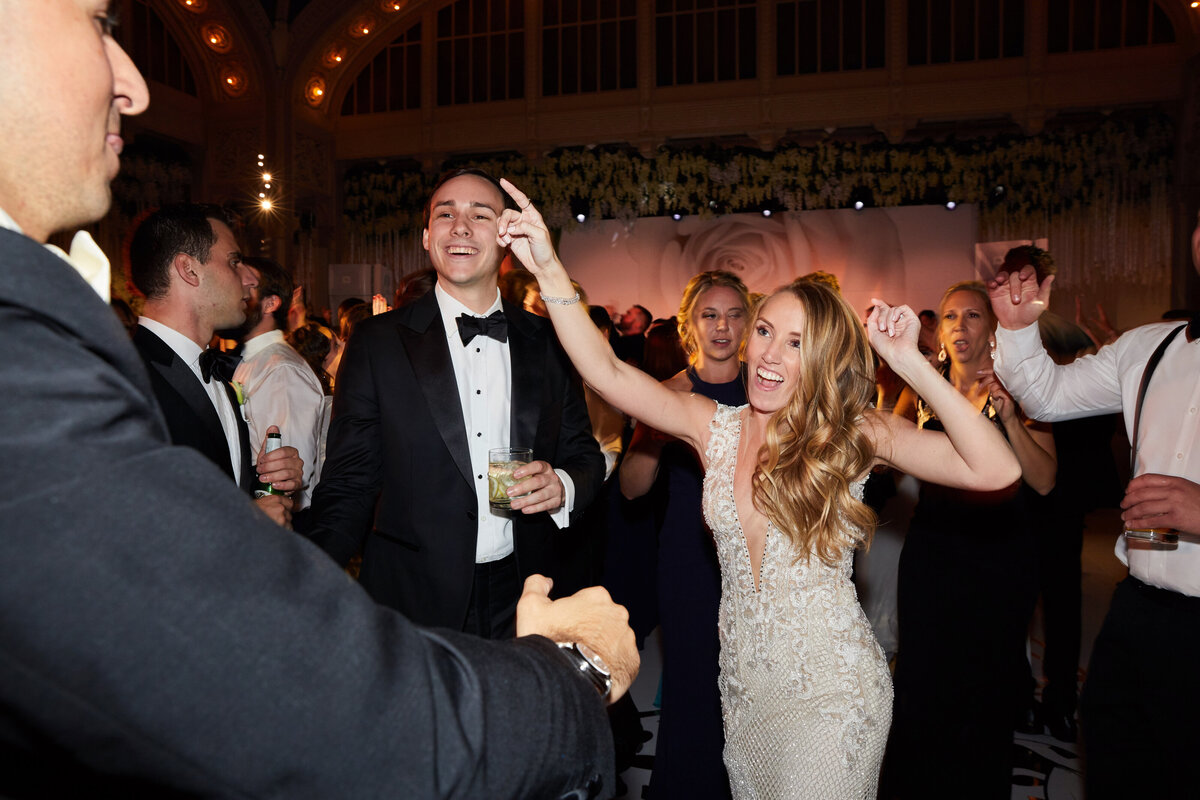 In the joyful photograph captured by photographer Christian Oth at The New York Public Library in New York City, a radiant bride in a stunning white dress can be seen energetically dancing with a beaming groom in a dashing tuxedo. The lively wedding reception setting is elegantly decorated, and the couple is surrounded by guests in formal attire who are clapping and enthusiastically joining in the celebration. East West Films, videographers for the event, beautifully captured these precious moments on video, adding an extra layer of joy and memories to the occasion.