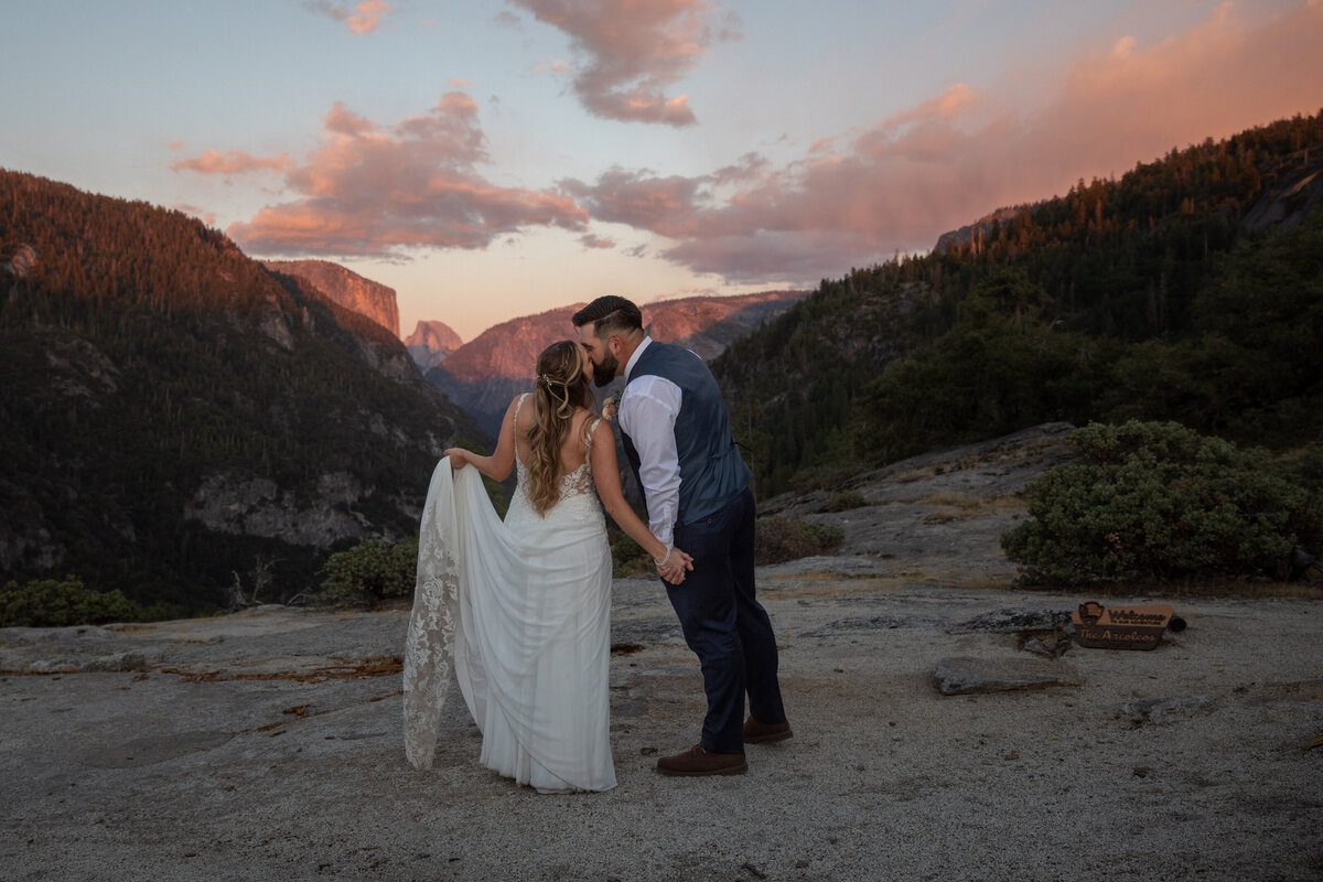 A bride and groom stand holding hands and kissing as the bride holds up the bottom of her wedding dress in Yosemite.
