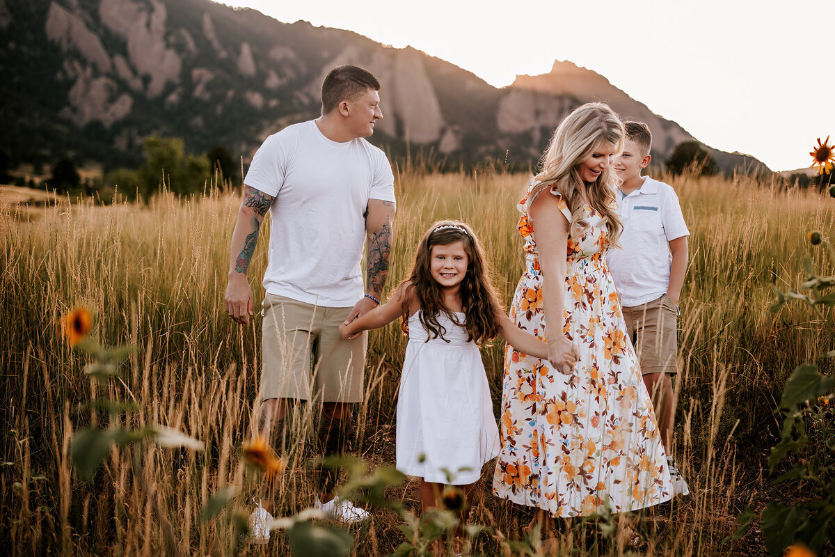 Family of four holding hands while walking through tall grass with the Boulder Flatirons in the background