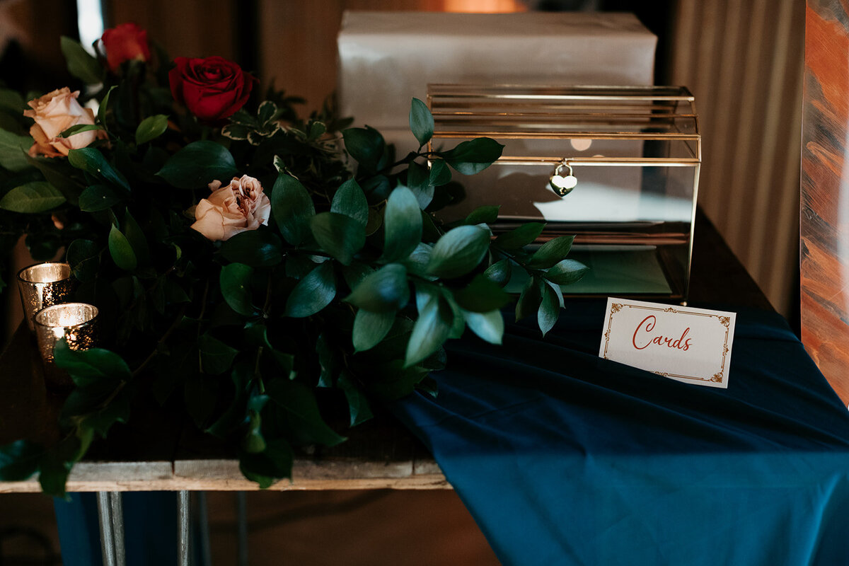 Card table display with a clear box labeled “Cards” beside floral greenery and small candles, creating an elegant and warm setup.