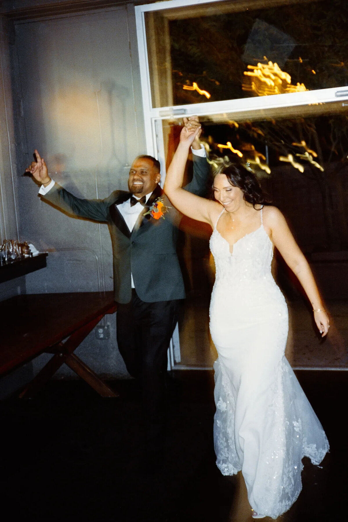 A smiling bride in a white lace gown and a groom in a dark suit with an orange boutonnière enter hand in hand, celebrating with joyful expressions and raised arms—captured perfectly by an experienced NJ wedding photographer.