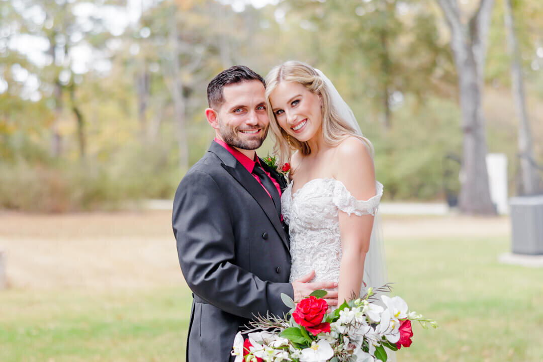 A bride and her groom in a grey and red suit snuggle in a lawn while smiling