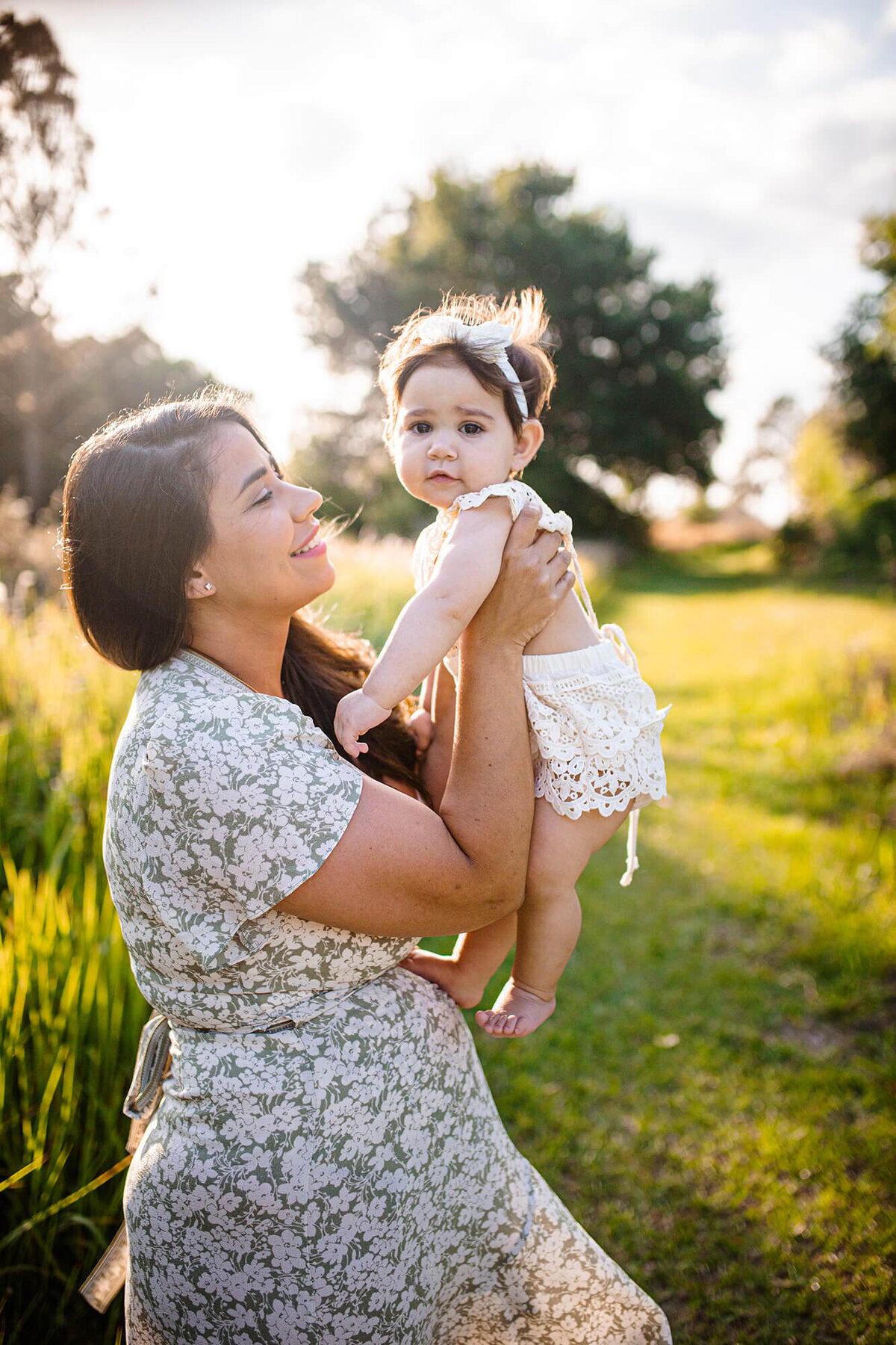 Sunset golden light of mother looking at daughter while holding her up in a field of tall grass. Captured at Alderman Ford Preserve by Sarah Wyndham Photography