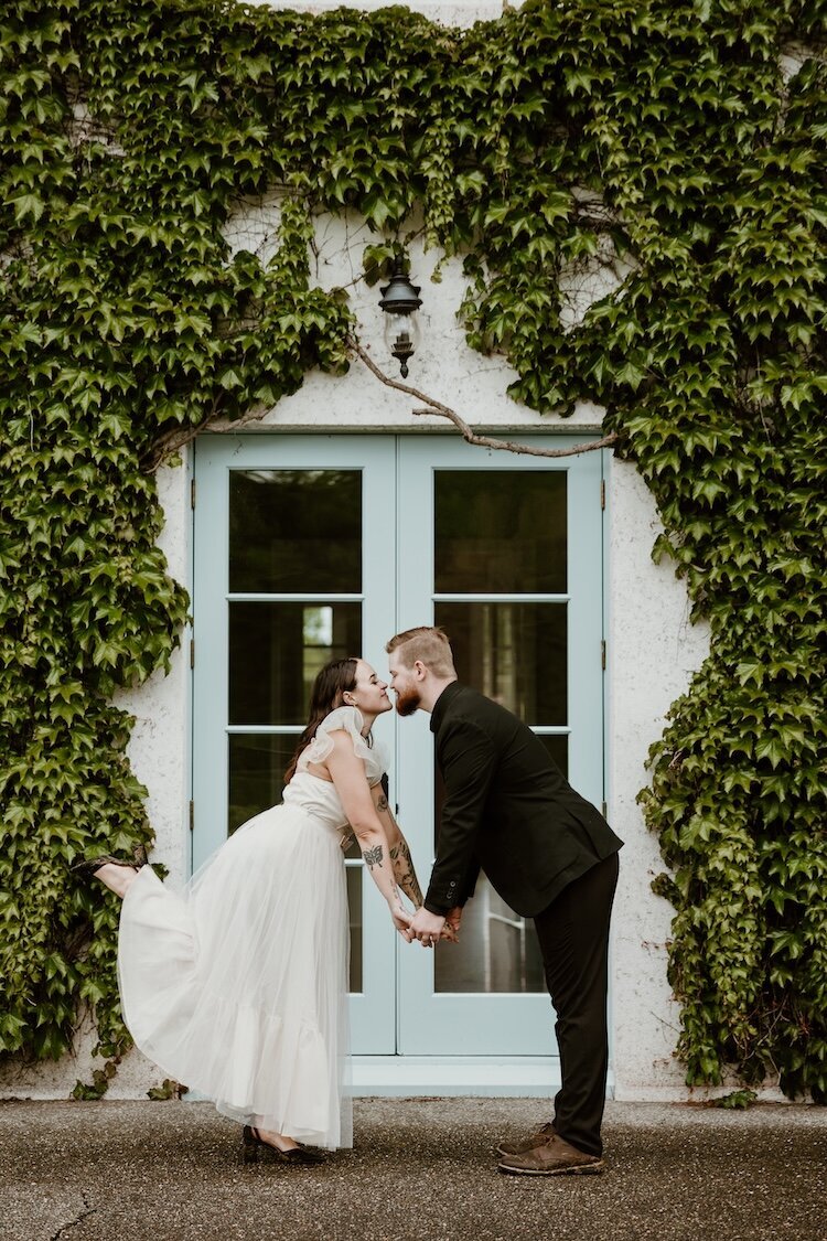 A happy engaged couple kisses and pops a leg in front of an ivy covered wall