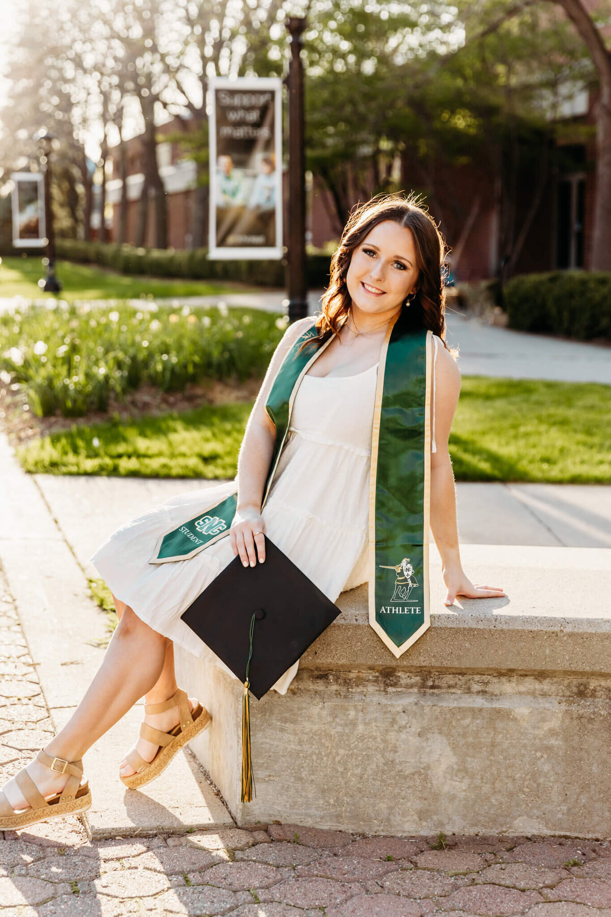 gorgeous college grad glowing as she sits on a cement bench and smiles while holding her graduation cap at sunset in Green Bay