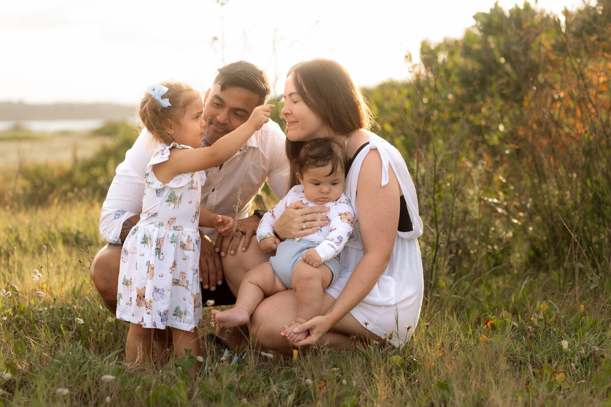 Young family sharing a tender moment in a sunlit grassy field, with a little girl gently touching her mother’s face while the father and baby look on lovingly.