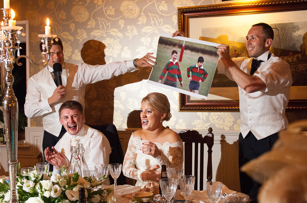 Bride in lace, princess wedding dress and blonde hair, laughing with groom during wedding speech by bestman at Ballyseede Castle