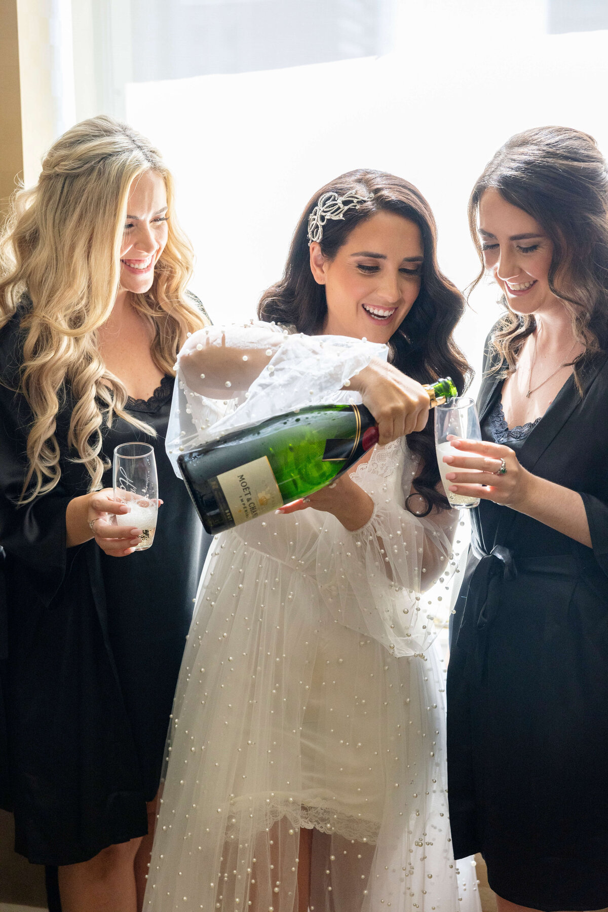 Two women in black robes share a joyful moment as the woman in the center, dressed in a white robe with pearl details, pours champagne into glasses. They stand by a window, smiling and celebrating.