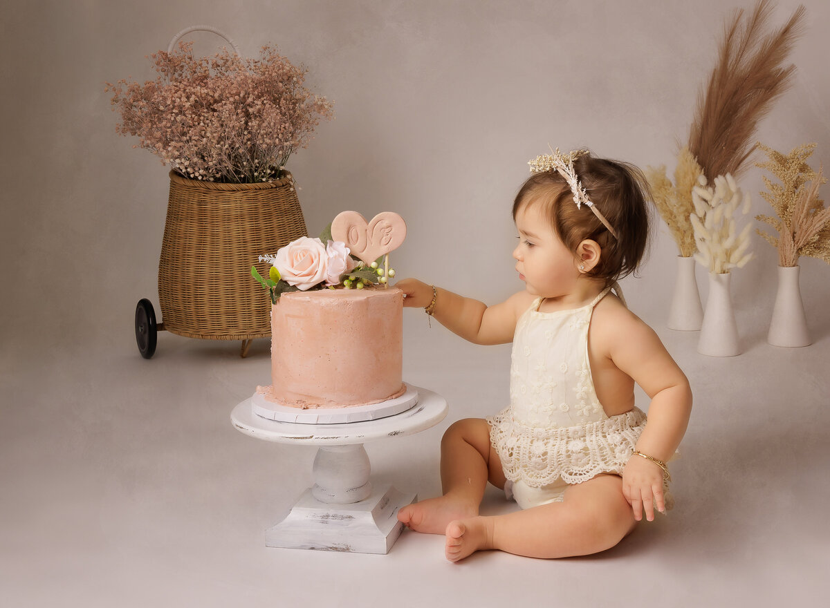 Baby girl cake smash photoshoot. Baby girl in white knit romper touches the top of her pink cake. She is wearing a delicate bow headband. Neutral blush photography set. Captured by best Brooklyn, NY cake smash photographer Chaya Bornstein.