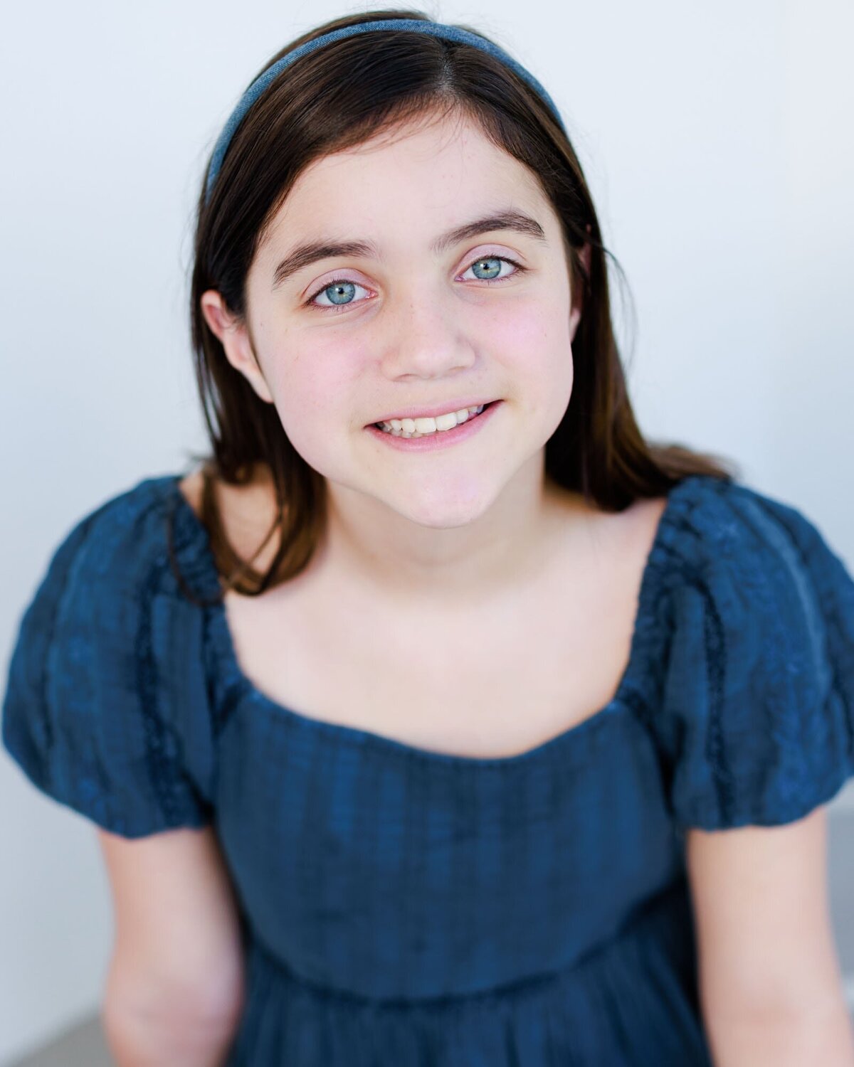 Actor headshot of a young girl wearing a blue dress on a white background.