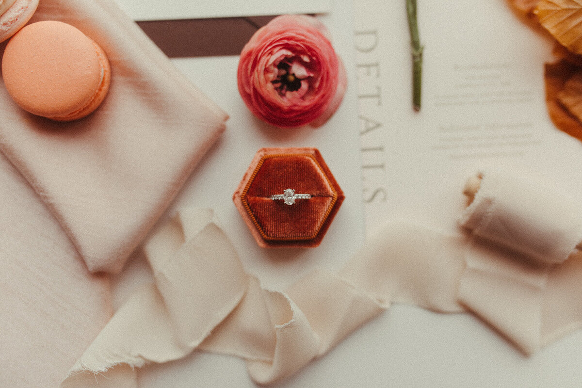 A wedding band in a velvet box, with flowers and a peach macaroon atop ribbon, and white wedding stationery with gray font.