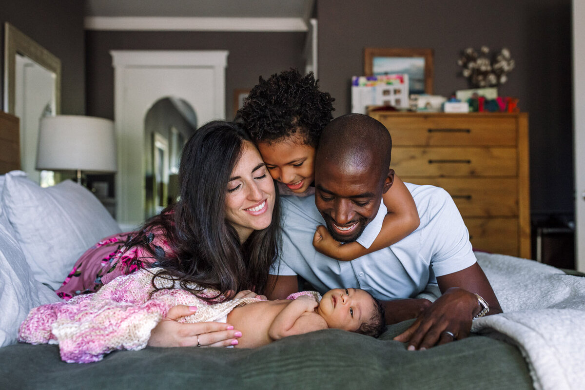 mom, dad, and 5-year-old boy on bed looking over at newborn sister