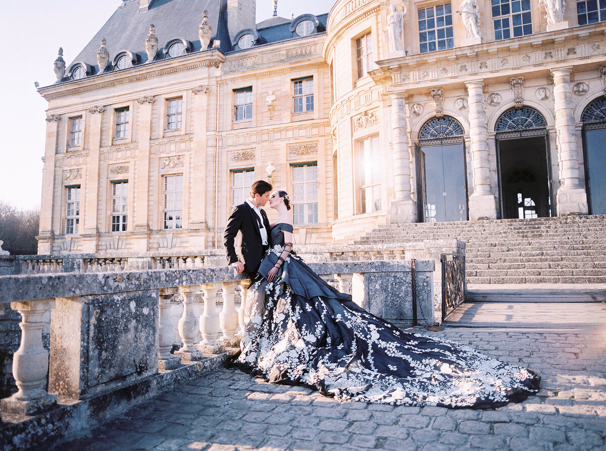 This stunning photo was taken at Chateau Vaux le Vicomte in Paris, France - known as one of the top destination wedding venues in the country. Captured by photographer Audrey, with videographers East West Films, the image showcases a couple standing on an outdoor stone terrace in front of an elegant, large building with ornate architecture. The woman looks exquisite in a black gown adorned with white floral details and a long train, while the man exudes sophistication in a black suit. Their affectionate gaze towards each other beautifully captures the essence of their special moment.