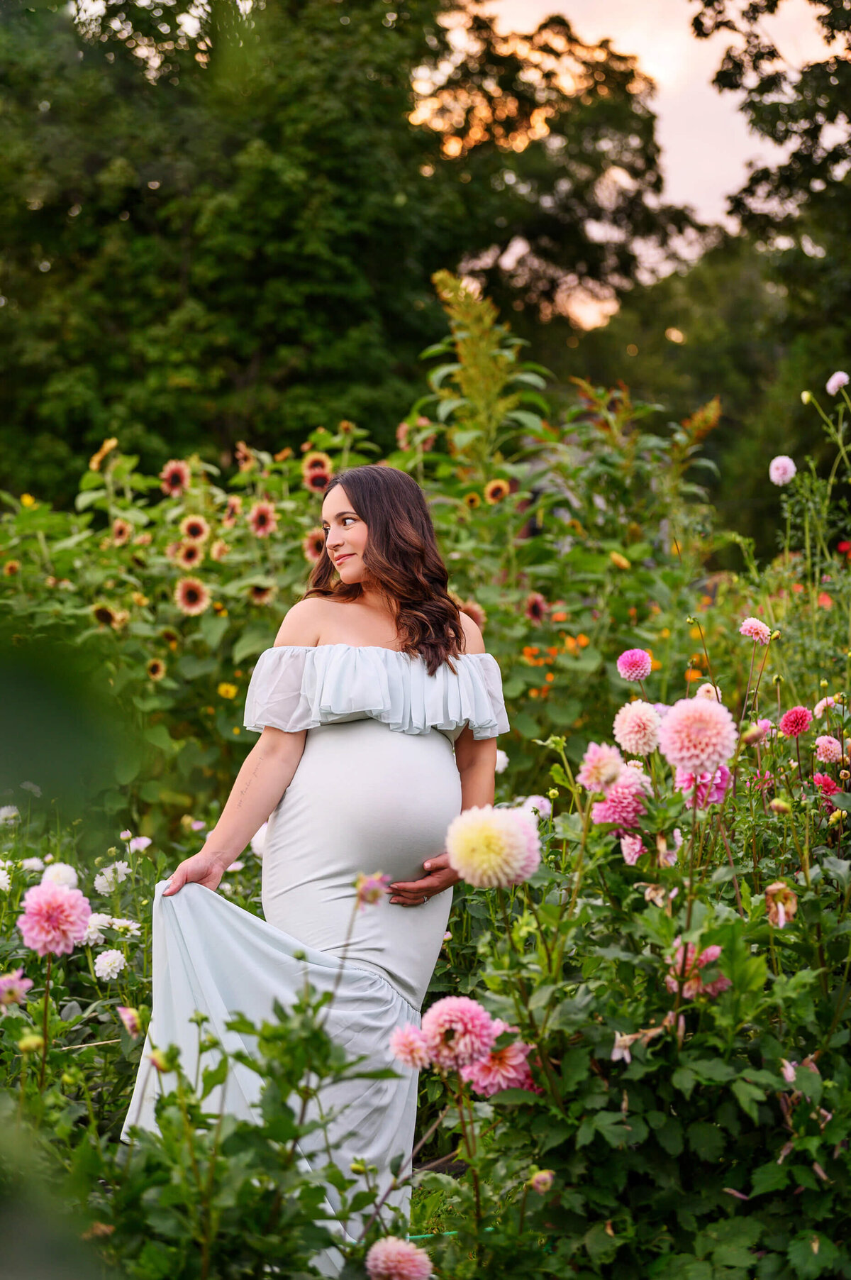 Pregnant woman wearing a mint green maternity dress standing in a flower field at sunset for her outdoor maternity photos in Burlington