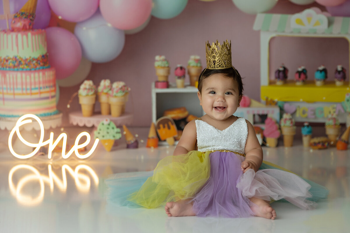 A happy one year old girl in a colorful dress and gold crown sits on the floor of her ice cream decorated birthday set