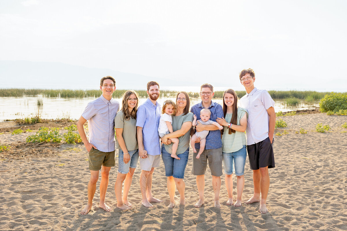 A large extended family poses barefoot on a sandy beach with a lake and mountains in the background. They are dressed in casual pastel and neutral tones, smiling warmly in the golden evening light.