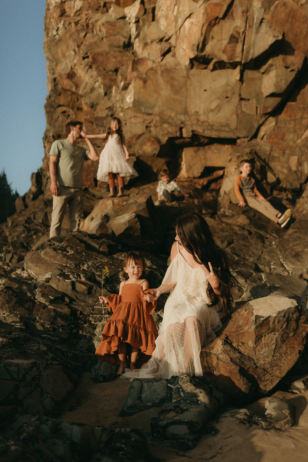 Lifestyle family portrait on rocky coastline Dad kisses daughter's hand, mom looks at daughter, brothers sit on rocks