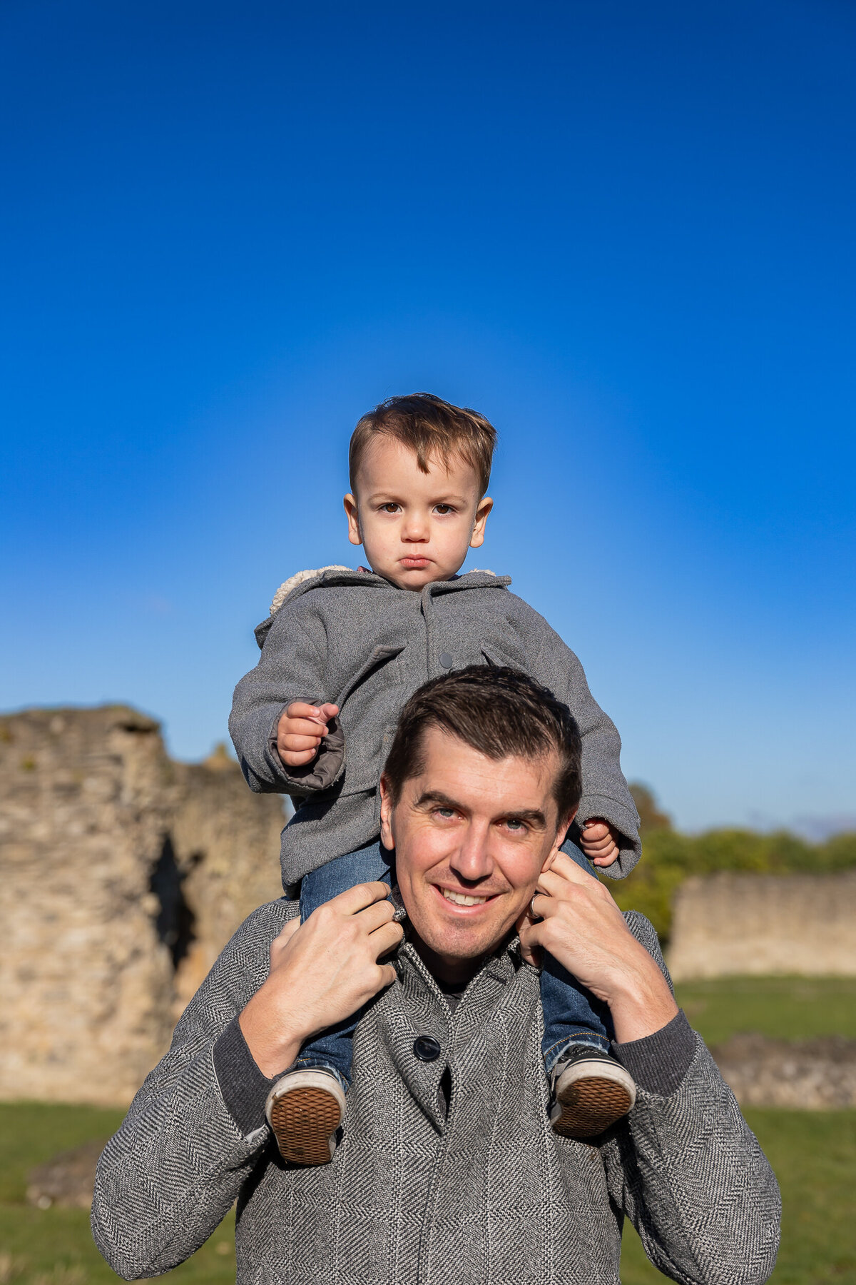 A young child sits on the shoulders of a smiling man outdoors with clear blue skies and old stone ruins in the background.